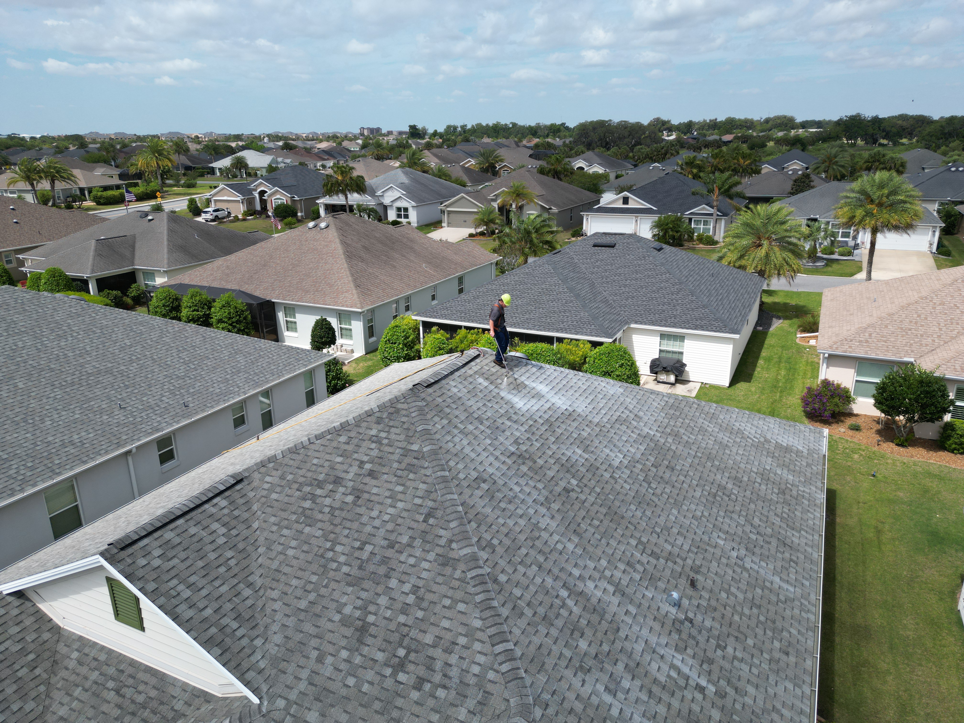Aerial view of a gray asphalt shingle roof being treated, with a technician on the roof in a neighborhood with palm trees